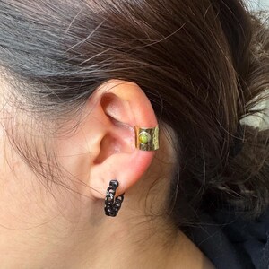May include: A gold-colored ear cuff with a textured surface and a small, round, green stone. A black chain-link hoop earring is also visible. The jewelry is worn on an ear.