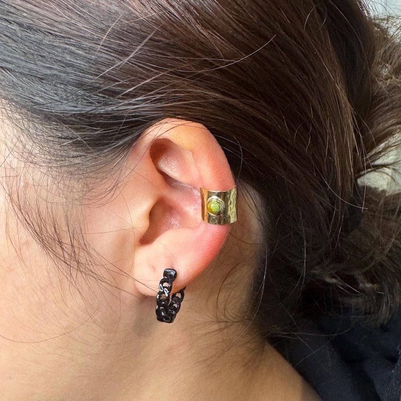 May include: A gold-colored ear cuff with a textured surface and a small, round, green stone. A black chain-link hoop earring is also visible. The jewelry is worn on an ear.