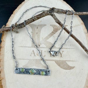 May include: A silver-toned necklace featuring a rectangular bar pendant with an etched pattern, set with two round green stones and one central square green stone. The necklace includes a delicate chain and a lobster claw clasp.