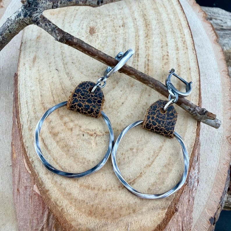 May include: A pair of silver hoop earrings with a hammered texture. Each earring features a small, dark brown leather accent at the top. The earrings are displayed on a wooden surface with a small branch.