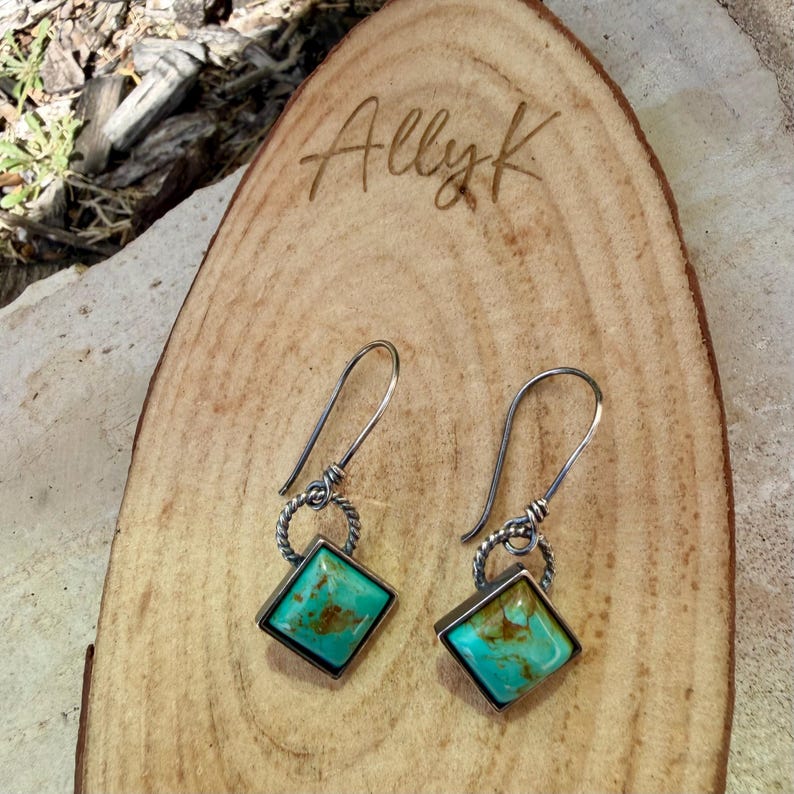 May include: A pair of turquoise and silver earrings. Each earring features a square turquoise stone set in a silver frame, with a twisted silver ring and a curved hook. The earrings rest on a wooden surface with the engraved text "Allyk".