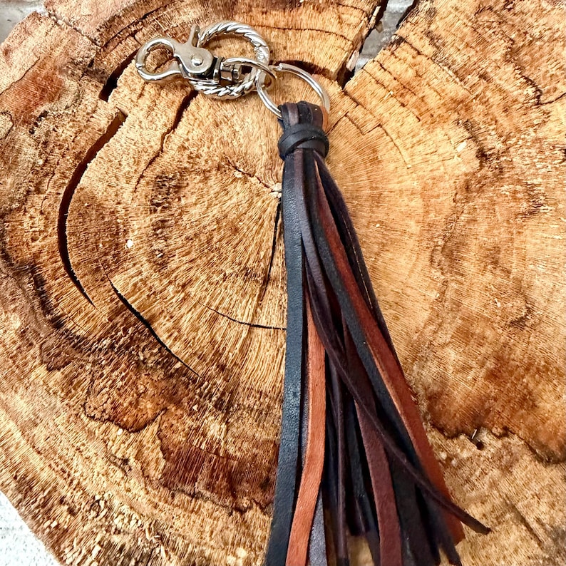 May include: A keychain with a silver clasp and ring, featuring a black, brown, and tan leather tassel. The keychain is displayed on a rustic, textured wooden surface, showcasing the natural wood grain.