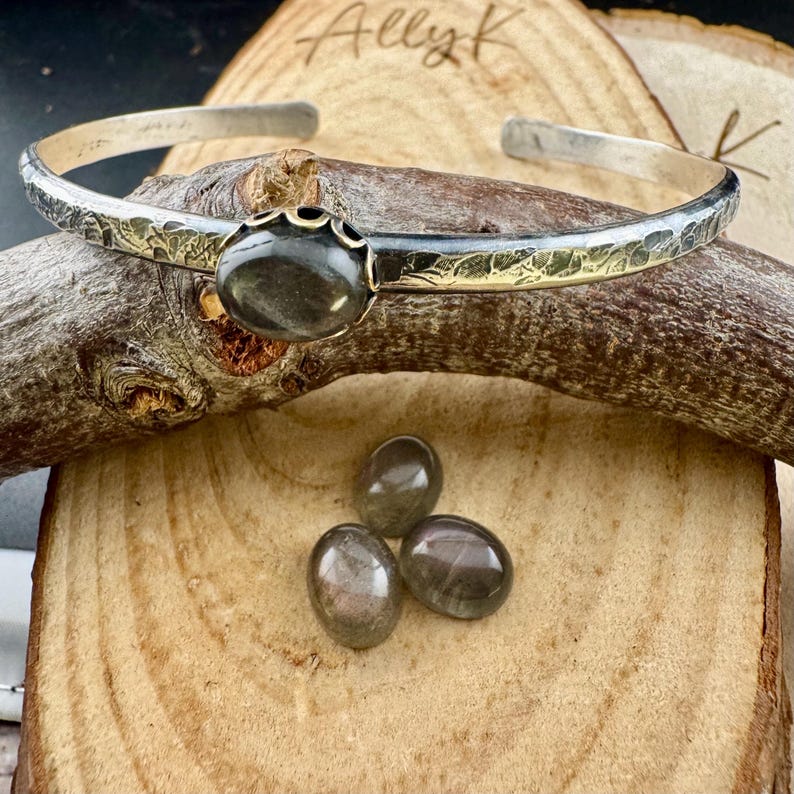 May include: A silver cuff bracelet with a textured gold band and a large, oval, dark gray gemstone. Three smaller, similar gemstones are displayed below the bracelet on a wooden surface. The name "Ally K" is handwritten in the upper left corner.