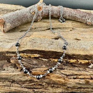 May include: A silver-toned necklace with a chain and black beads. The necklace is displayed on a piece of wood. The beads are evenly spaced along the necklace, with smaller silver beads between them. The necklace has a lobster clasp.