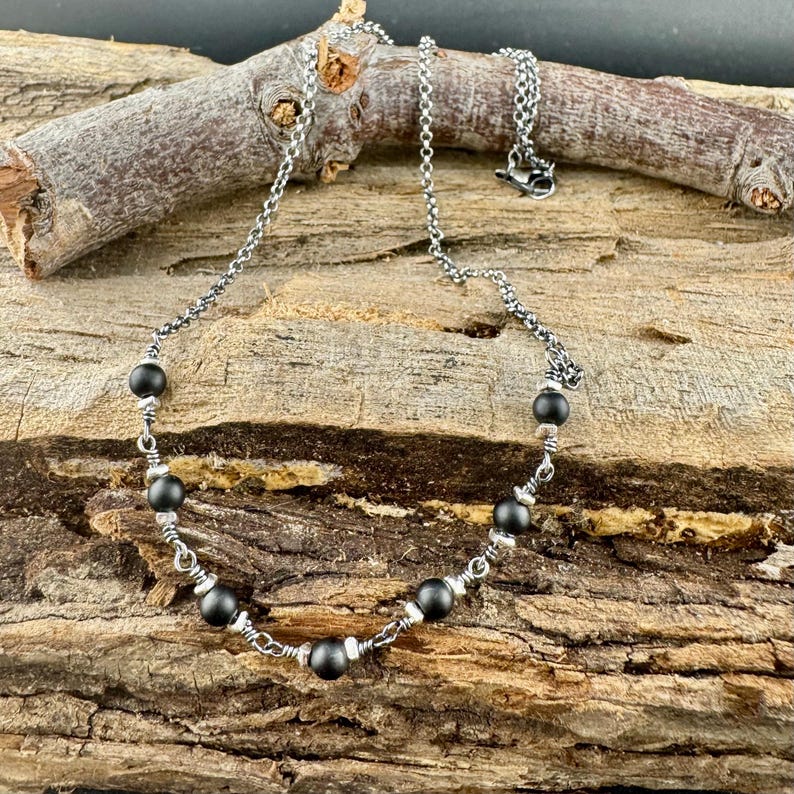 May include: A silver-toned necklace with a chain and black beads. The necklace is displayed on a piece of wood. The beads are evenly spaced along the necklace, with smaller silver beads between them. The necklace has a lobster clasp.
