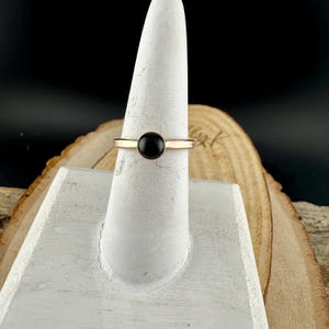 May include: A gold-coloured ring featuring a circular, black gemstone at its centre. The ring is presented on a white, conical stand, set against a wooden backdrop. The ring's band is slender and plain, with a polished surface.