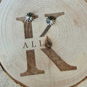 May include: Two silver-toned stud earrings, each featuring a sunburst or star-like design with radiating points. The earrings are placed on a light-colored wooden slice engraved with a large letter K and the text ALL.