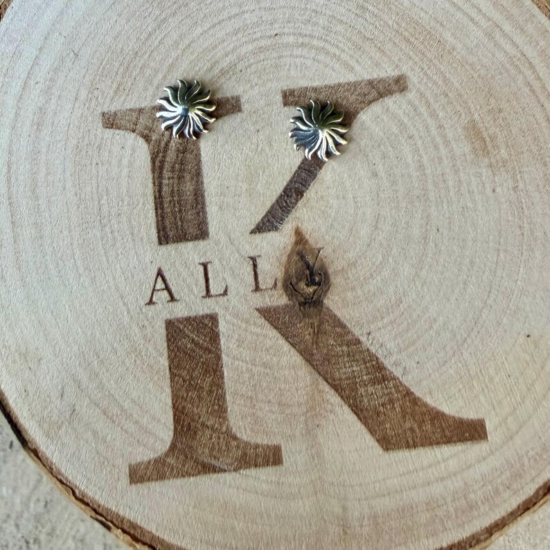 May include: Two silver-toned stud earrings, each featuring a sunburst or star-like design with radiating points. The earrings are placed on a light-colored wooden slice engraved with a large letter K and the text ALL.