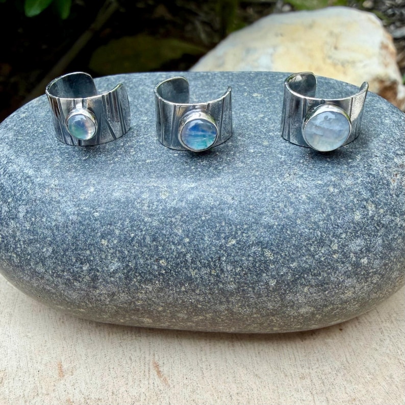 K&ouml;nnte beinhalten: Drei silberfarbene, verstellbare Ringe mit strukturiertem Band und einem runden, irisierenden Stein. Die Ringe werden auf einem grauen Stein pr&auml;sentiert, der ihr Design hervorhebt. Die Steine weisen blaue, wei&szlig;e und gr&uuml;ne Farbt&ouml;ne auf.