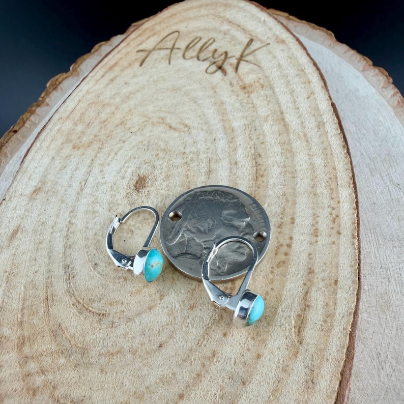 May include: Silver leverback earrings with turquoise stones, displayed on a wooden surface alongside a coin. The earrings have a simple design. The wooden surface has the inscription "Ally K".