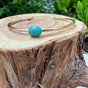 May include: A gold-colored cuff bracelet with a round, turquoise stone set in the center. The bracelet is displayed on a rustic, wooden surface, with green foliage in the background. The bracelet has a simple, elegant design.