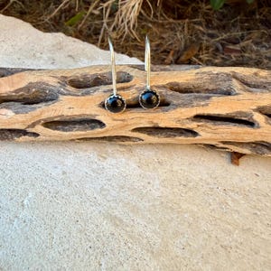 May include: A pair of silver drop earrings featuring round, polished black stones set in scalloped metal bezels. The earrings have curved silver wires that extend upward from the stone settings, creating a simple and clean design.