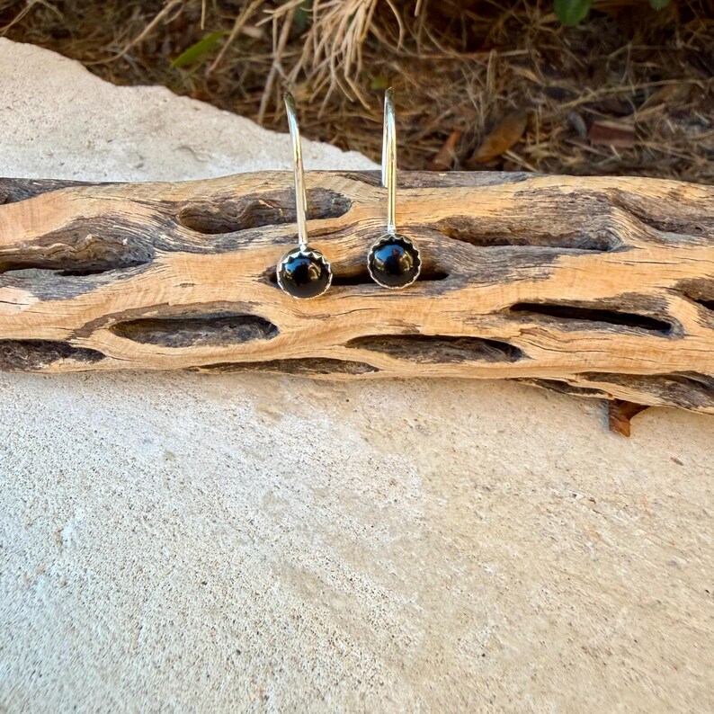 May include: A pair of silver drop earrings featuring round, polished black stones set in scalloped metal bezels. The earrings have curved silver wires that extend upward from the stone settings, creating a simple and clean design.