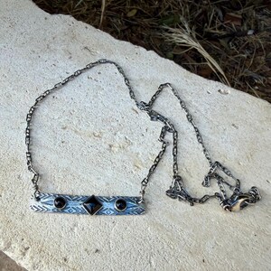 Pu&ograve; includere: Una collana a barra in metallo color argento con una pietra nera centrale a forma di diamante affiancata da due pietre nere rotonde pi&ugrave; piccole. La barra presenta un motivo geometrico inciso ed &egrave; appesa a una catena a maglie rettangolari.