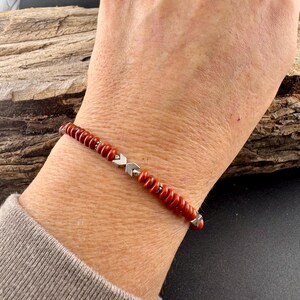 May include: A red beaded bracelet with silver-colored accents. The bracelet features small, round, red beads strung together with silver-toned metal pieces. The bracelet is worn on a wrist, showcasing its design and color.