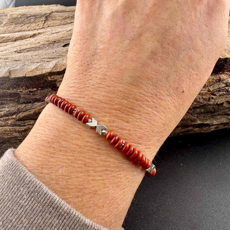 May include: A red beaded bracelet with silver-colored accents. The bracelet features small, round, red beads strung together with silver-toned metal pieces. The bracelet is worn on a wrist, showcasing its design and color.