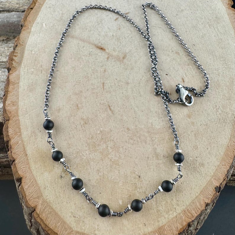 May include: A silver necklace with black beads. The necklace features a delicate chain with evenly spaced, matte black beads. The beads are interspersed with small silver accents, creating a contrast. The necklace is displayed on a wooden surface.