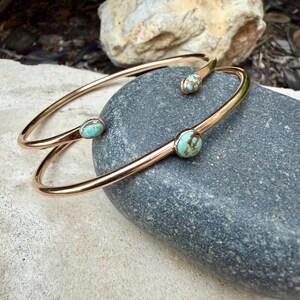 May include: Two open-ended, gold-colored bangles with turquoise-colored stone accents. The bangles are resting on a gray stone surface. The turquoise stones have brown veining. The bangles are a simple, elegant design.