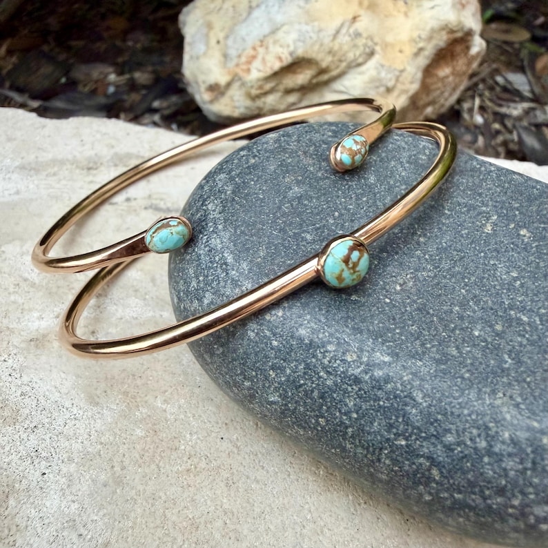 May include: Two open-ended, gold-colored bangles with turquoise-colored stone accents. The bangles are resting on a gray stone surface. The turquoise stones have brown veining. The bangles are a simple, elegant design.