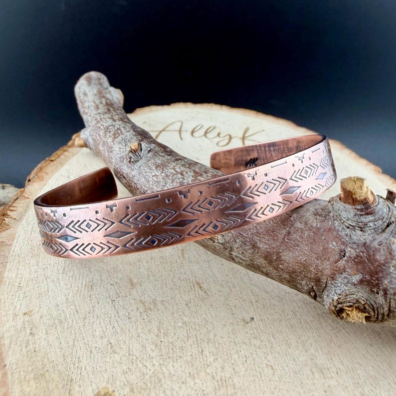 May include: A copper cuff bracelet featuring a repeating geometric pattern of etched diamonds and chevrons. The metal has a warm, reddish-brown tone with darkened recessed lines. The interior of the band is stamped with the text Ally K.