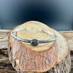 May include: A silver cuff bracelet with a dark, oval-shaped stone at the center. The bracelet has a textured, floral design along the band. The bracelet is displayed on a wooden surface.