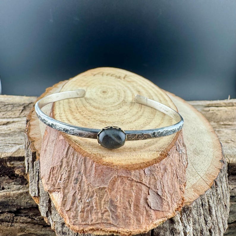 May include: A silver cuff bracelet with a dark, oval-shaped stone at the center. The bracelet has a textured, floral design along the band. The bracelet is displayed on a wooden surface.