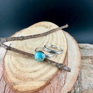 May include: Two silver rings displayed on a wooden surface. One ring features a round, turquoise stone. The other ring has a textured band. A small twig rests across the rings, adding a natural element to the composition.