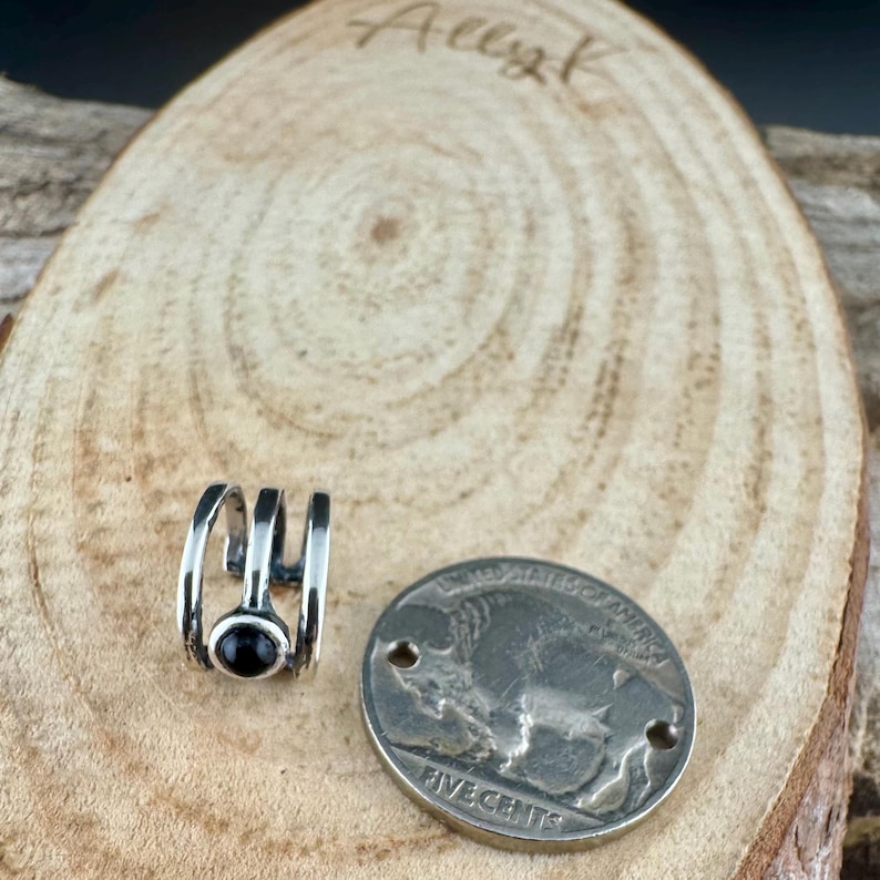 May include: A silver-toned ear cuff with a black stone, next to a silver coin. The ear cuff has a three-bar design. The coin has a buffalo image and the words "FIVE CENTS". The background is a wooden surface.