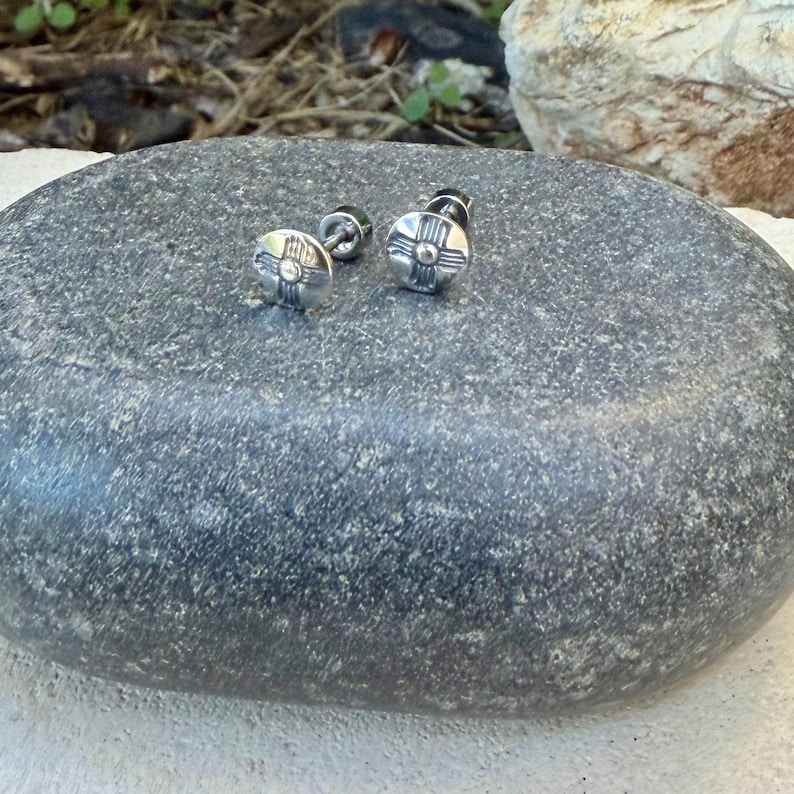 May include: A pair of circular silver stud earrings featuring a raised central dot surrounded by four radiating lines, creating a cross-like pattern. The earrings have a polished metallic finish and are designed with a post-back closure mechanism.