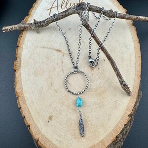 May include: A silver-toned necklace with a turquoise teardrop-shaped stone and a feather pendant. The necklace features a twisted circle design and is displayed on a wooden surface with a small branch.