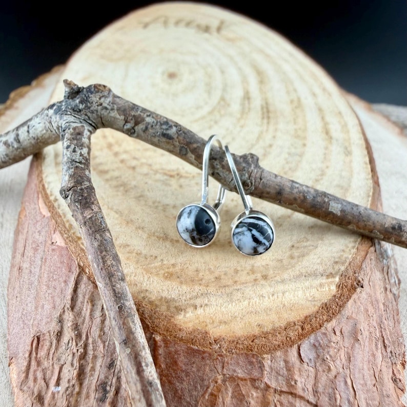 May include: A pair of silver drop earrings with round, black and white patterned stones. The earrings are displayed on a small wooden branch, set against a textured wooden background.