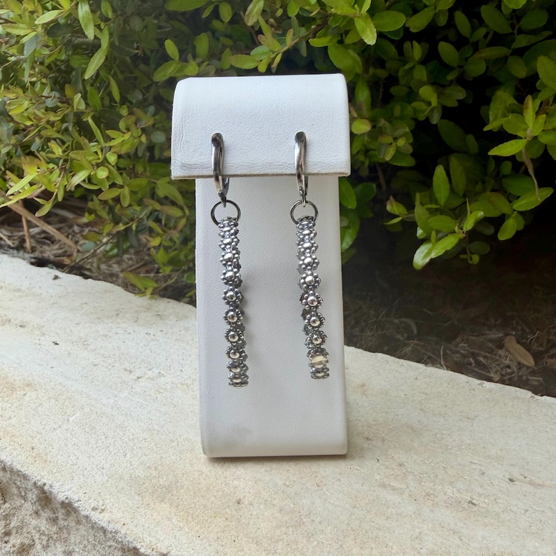 May include: Silver-toned dangle earrings with a floral design. The earrings are displayed on a white stand against a backdrop of green foliage. The earrings have a hoop closure and a long, beaded design.