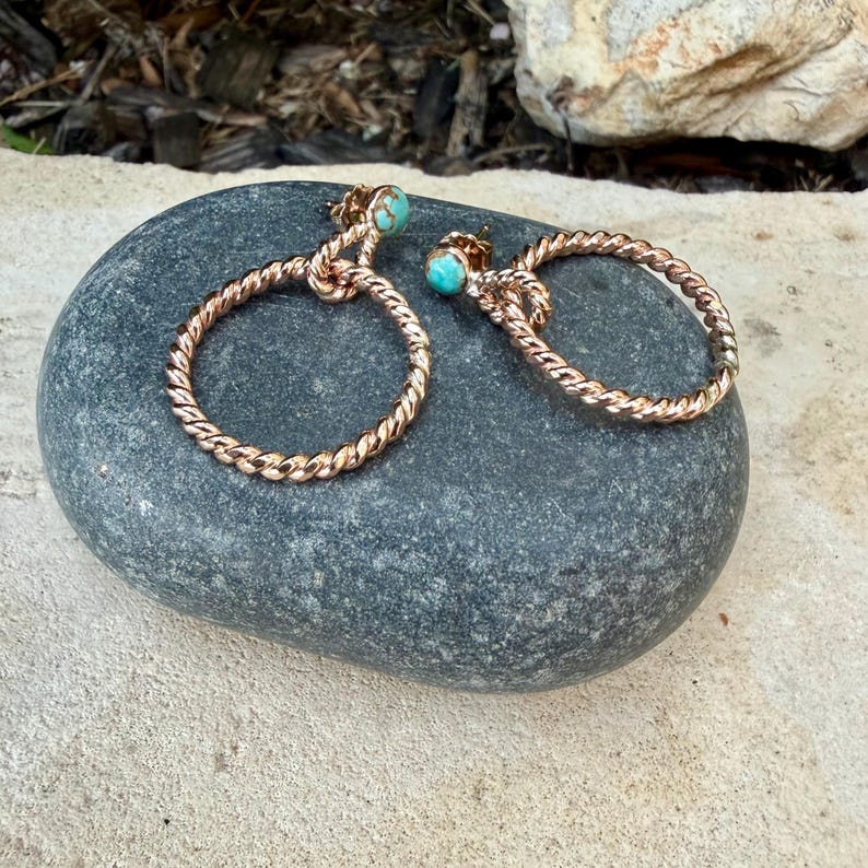 May include: A pair of copper-colored hoop earrings with a twisted rope design. Each earring features a small, round, turquoise-colored stone at the top. The earrings are displayed on a smooth, gray stone surface.