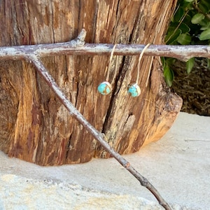 May include: A pair of dangle earrings featuring round, polished turquoise stones set in gold-toned metal. Each earring hangs from a curved, slender gold-toned wire hook, showcasing a smooth, vibrant blue-green surface with natural dark veining.