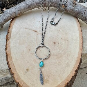 May include: A silver-toned necklace featuring a circular pendant with a twisted design. A turquoise-colored stone and a small feather charm dangle from the circle. The necklace rests on a wooden surface.