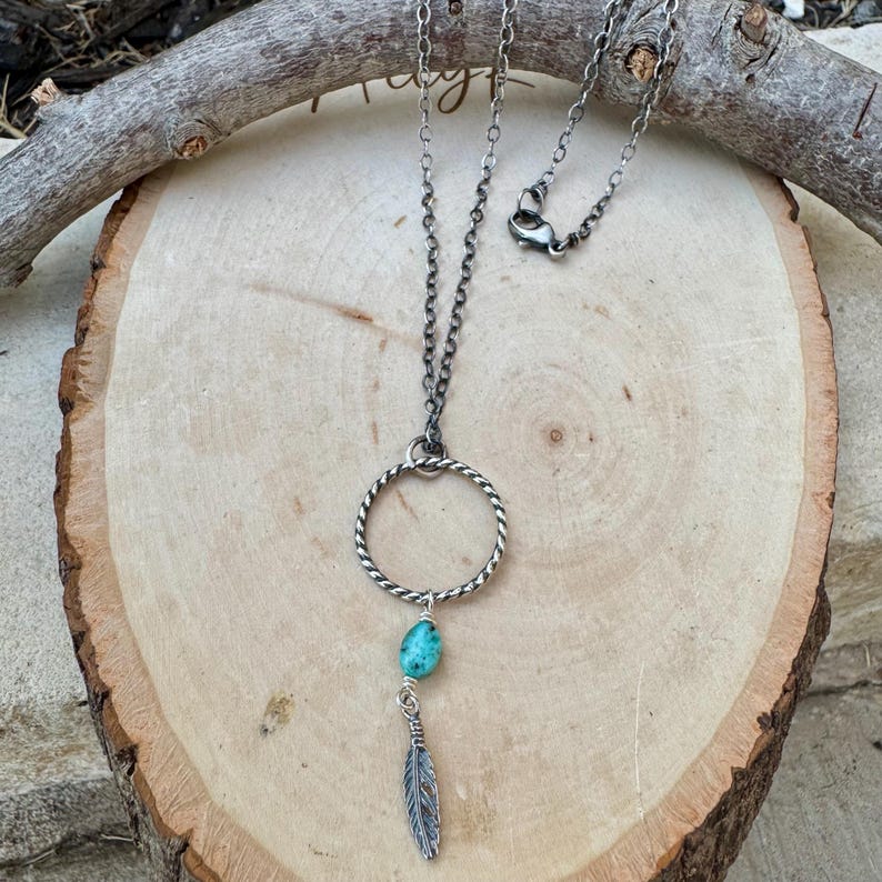 May include: A silver-toned necklace featuring a circular pendant with a twisted design. A turquoise-colored stone and a small feather charm dangle from the circle. The necklace rests on a wooden surface.