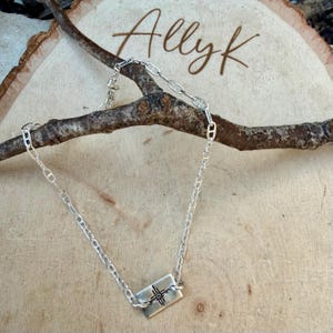 May include: A silver chain necklace with a rectangular pendant featuring a cross design. The necklace is displayed on a wooden surface with the name "Allyk" written in cursive.
