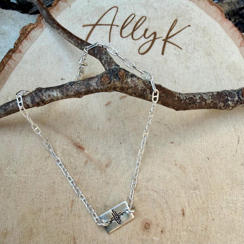 May include: A silver chain necklace with a rectangular pendant featuring a cross design. The necklace is displayed on a wooden surface with the name "Allyk" written in cursive.
