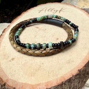 May include: A handcrafted bracelet featuring a braided leather band in shades of brown and tan, intertwined with a strand of black beads and turquoise-colored stones. The bracelet is displayed on a wooden surface, showcasing a rustic aesthetic.