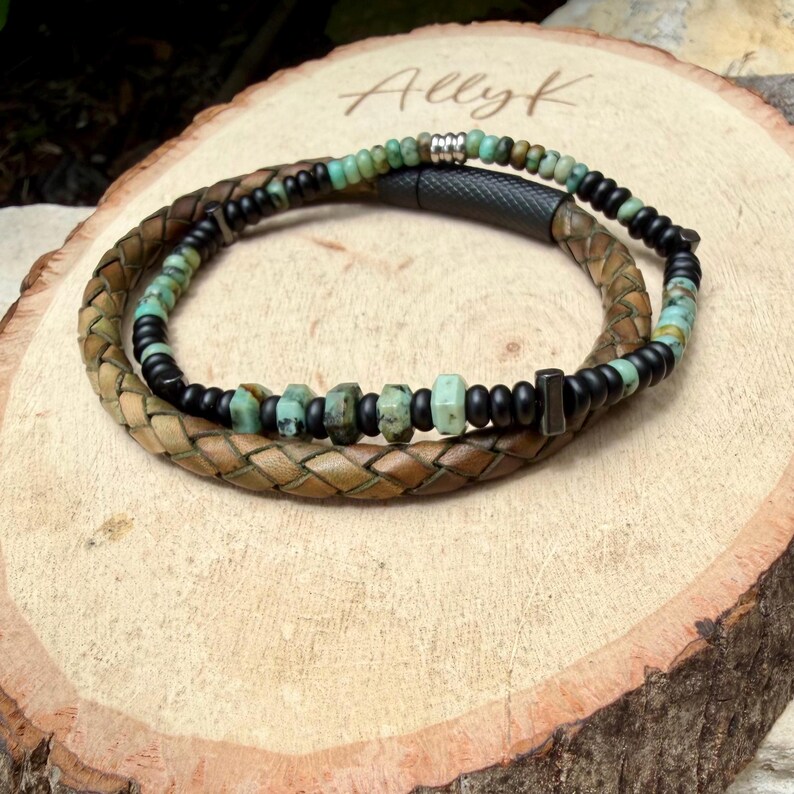 May include: A handcrafted bracelet featuring a braided leather band in shades of brown and tan, intertwined with a strand of black beads and turquoise-colored stones. The bracelet is displayed on a wooden surface, showcasing a rustic aesthetic.