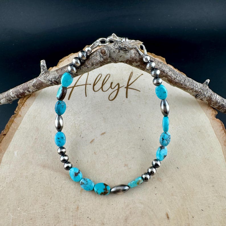 May include: Turquoise and silver beaded bracelet. The bracelet features oval turquoise stones and silver beads. The bracelet is strung on a silver chain and is displayed on a wooden surface.