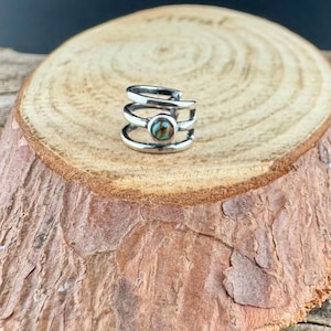 May include: A silver ear cuff with a turquoise stone set in the center. The cuff wraps around the ear with three stacked bands. The jewelry is displayed on a wooden surface.