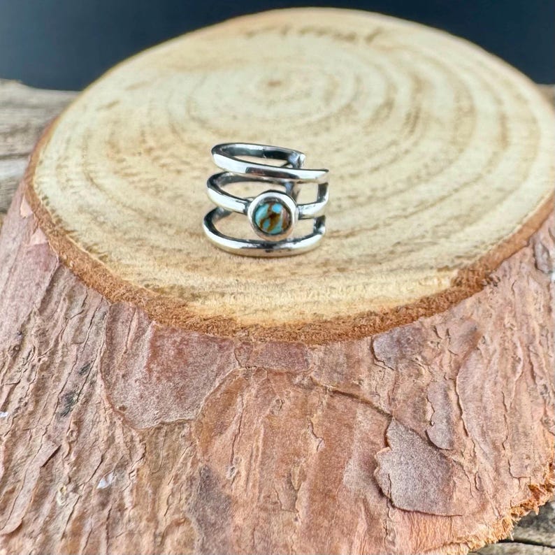May include: A silver ear cuff with a turquoise stone set in the center. The cuff wraps around the ear with three stacked bands. The jewelry is displayed on a wooden surface.