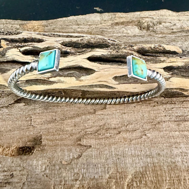 Op de afbeelding: Een zilverkleurige manchetarmband met een gedraaid touwontwerp. De armband heeft twee vierkante turkooizen stenen gezet in zilver. De armband wordt getoond op een stuk verweerd hout.