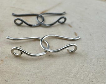 May include: Two silver-colored metal jewelry clasps. Each clasp has a curved hook shape with a looped end. One clasp is in focus, while the other is slightly out of focus in the background. The clasps are on a light-colored wooden surface.