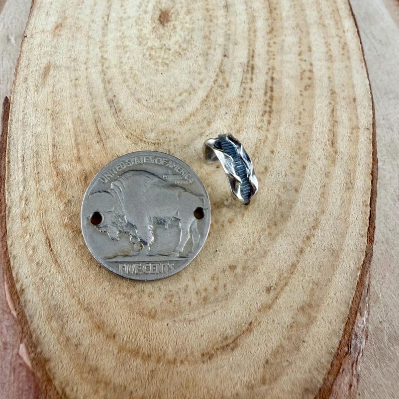 May include: A silver-toned ring with a textured design sits next to a silver buffalo nickel. The coin is stamped with the words "United States of America" and "Five Cents". The ring has a unique, organic shape.