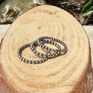 Puede incluir: Tres anillos plateados con cuentas se exhiben sobre una rodaja de madera natural. Los anillos est&aacute;n hechos de peque&ntilde;as cuentas redondas y est&aacute;n dispuestos de forma ligeramente superpuesta. La madera tiene un color marr&oacute;n claro con vetas visibles.