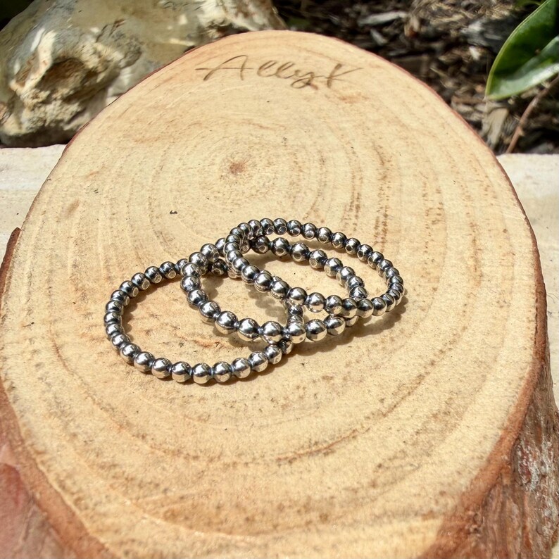 Puede incluir: Tres anillos plateados con cuentas se exhiben sobre una rodaja de madera natural. Los anillos est&aacute;n hechos de peque&ntilde;as cuentas redondas y est&aacute;n dispuestos de forma ligeramente superpuesta. La madera tiene un color marr&oacute;n claro con vetas visibles.