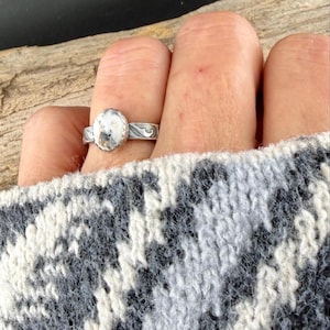 May include: A silver ring with an oval, white and brown stone is worn on a finger. The ring has a decorative, swirling design on the band. The person is wearing a gray and white patterned sweater.