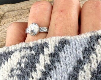 Op de afbeelding: Een zilveren ring met een ovale, wit en bruine steen wordt op een vinger gedragen. De ring heeft een decoratief, wervelend ontwerp op de band. De persoon draagt een grijs-witte trui met patroon.